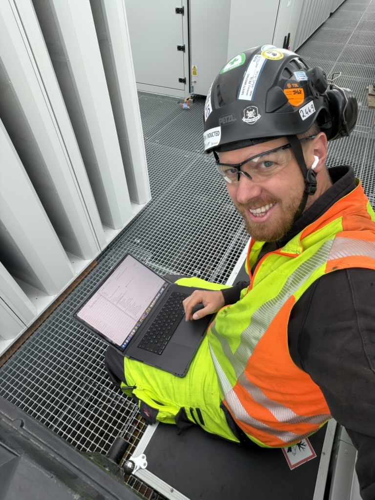 Tobias Schott von Omega Mounting dokumentiert einen Montageeinsatz am Laptop – mit Helm, Schutzbrille und Warnschutzkleidung auf dem Technikgang eines Rechenzentrums