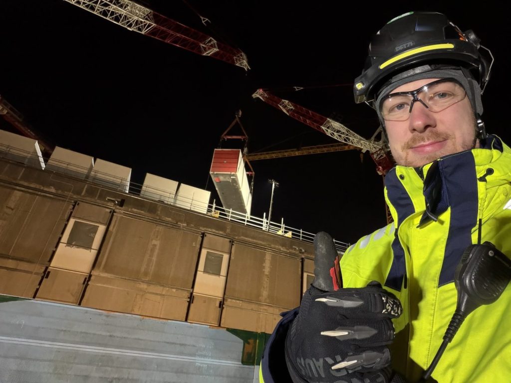Tobias Schott von Omega Mounting als Hebeaufsicht bei Nachtarbeit auf einer Industriebaustelle, im Hintergrund Turmdrehkrane und ein Container am Haken